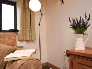 Ein Zimmer mit Fenster, einer Lampe, einer Couch mit einem Buch und einer Pflanze in einer weißen Vase.