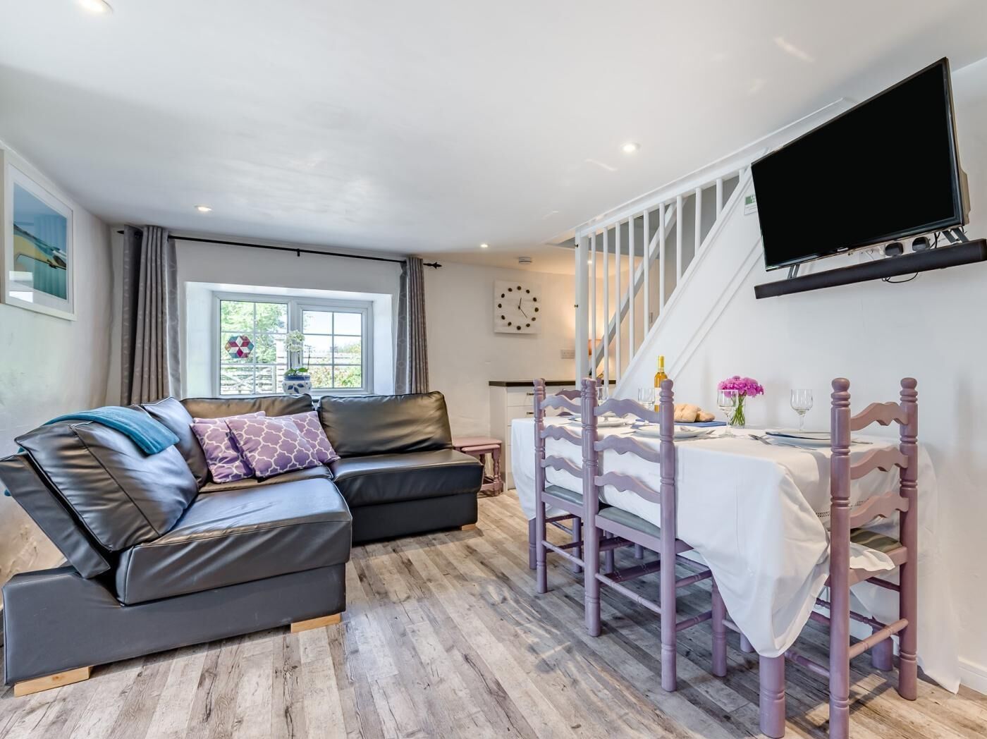 A modern room with a sofa, a dining table with chairs, a TV mounted on the wall, and a window with curtains.