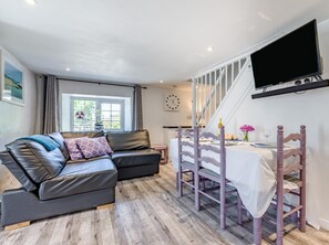 A modern room with a sofa, a dining table with chairs, a TV mounted on the wall, and a window with curtains. - 2 Bed in Bude (oc-00952) (Launcells)