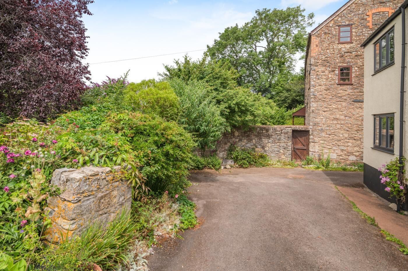 A stone building with a paved driveway leading to a wooden gate, surrounded by lush greenery and flowering plants.