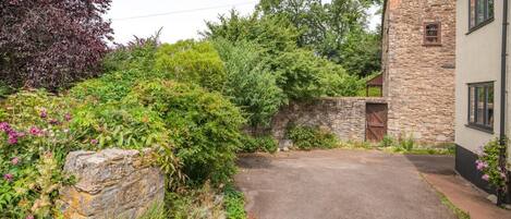 A stone building with a paved driveway leading to a wooden gate, surrounded by lush greenery and flowering plants.
