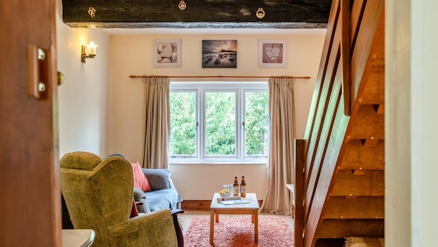 A cozy room with a sofa, armchair, and a small table with bottles. A window with curtains and a view of greenery is visible.