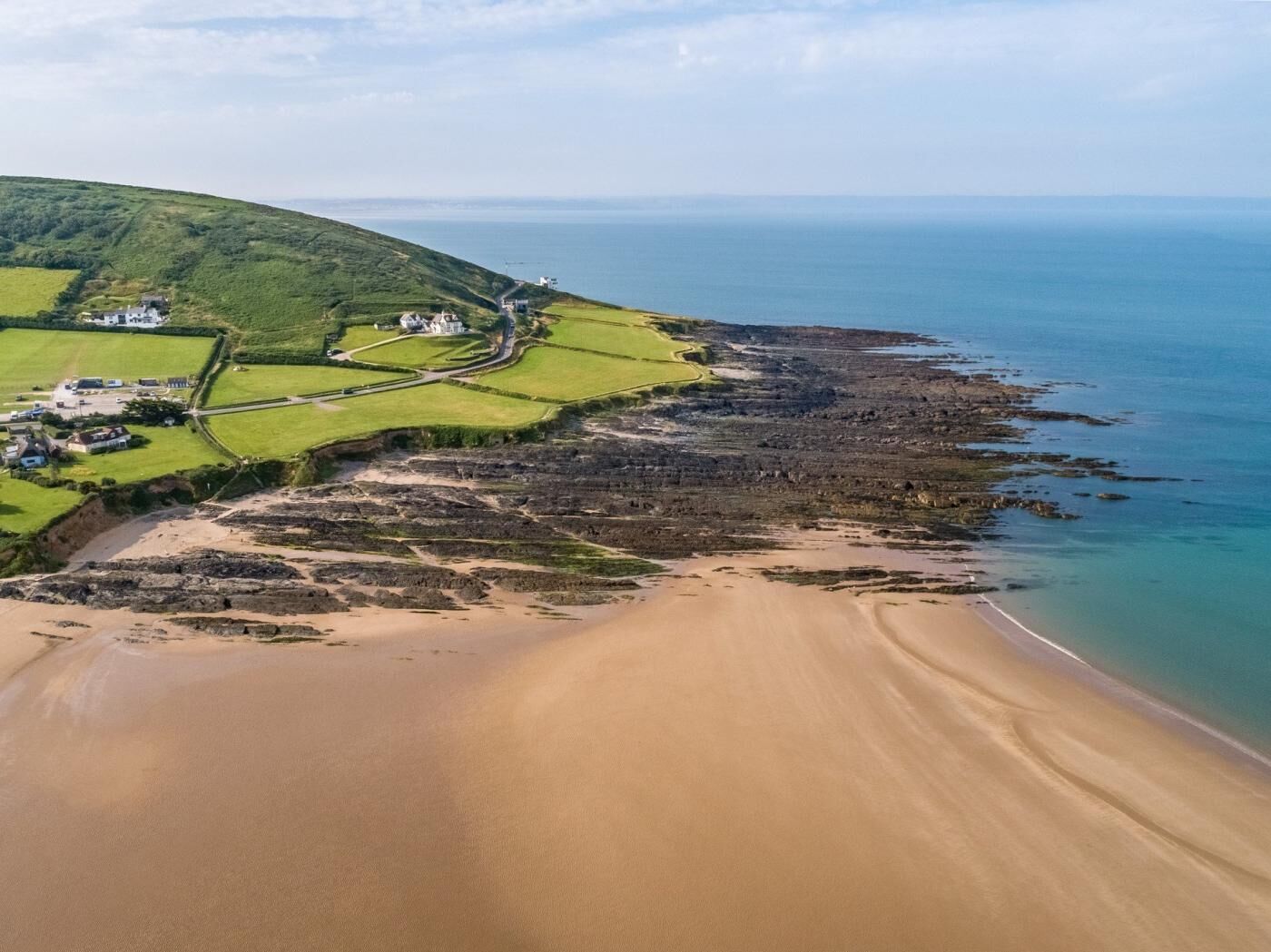 Une vue aérienne d’une zone côtière avec une plage de sable, des champs verdoyants et quelques bâtiments.