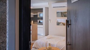 A hotel room with a bed, a round table with a book and a vase, a kitchenette in the background, and a door with a number sign. - Corporate Stays Insignia City - B52 (Asunción)