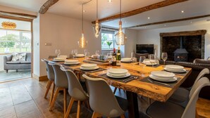 Un comedor con una larga mesa de madera dispuesta para seis personas, sillas grises, lámparas colgantes y una chimenea de piedra al fondo.