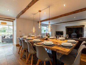 A dining area with a long wooden table set for six, grey chairs, hanging pendant lights, and a stone fireplace in the background. - 6 Bed in Bude (oc-oldfa) (Marhamchurch)