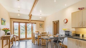 A spacious kitchen with wooden cabinets, a dining area with chairs, a table set for a meal, and a view through the glass doors. - 3 Bed in Callington (oc-mbarn) (Linkinhorne)
