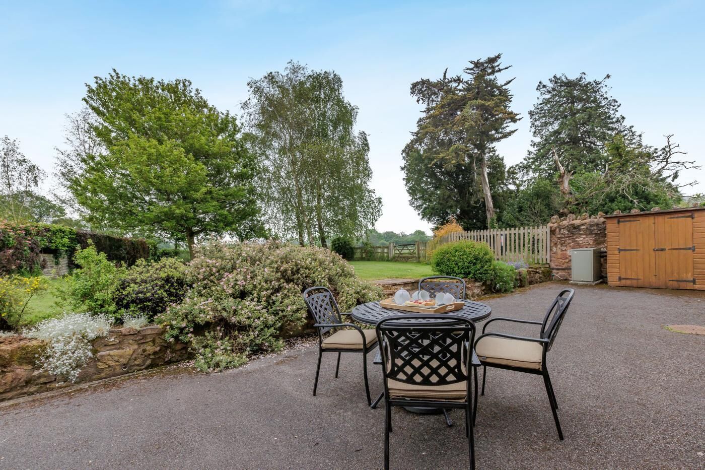 A patio with a round table and four chairs, surrounded by a garden with various plants and trees.