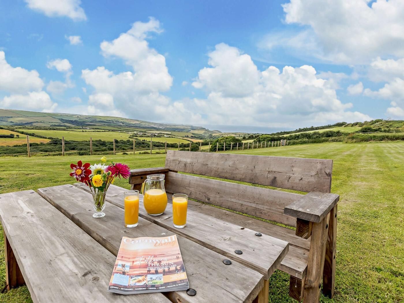 A wooden bench with a magazine, a vase with flowers, and two glasses of orange juice.