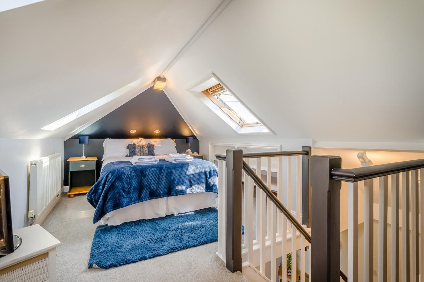 Ein Schlafzimmer mit schrägen Wänden, einem Bett mit blauer Decke, einem Nachttisch und einem Schreibtisch mit Lampe.