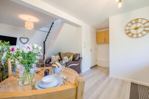 A dining area with a wooden table set for two, a staircase, a kitchen with a clock, and a living area with a sofa. - 1 Bed in Nr Bude (oc-penke) (Crackington)