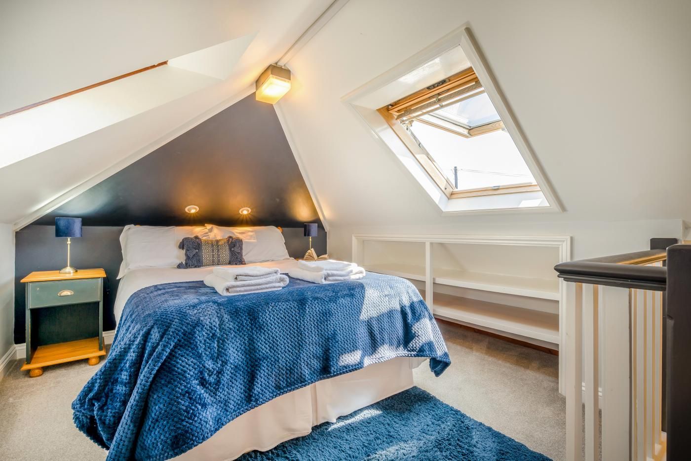 A bedroom with a sloped ceiling, a skylight, a bed with a blue blanket, a nightstand, and a bedside lamp.