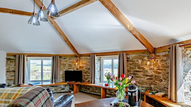 A cozy room with a stone wall, a wooden ceiling, a flat-screen TV, a dining table with a vase of flowers, and a sofa with a plaid blanket. - 1 Bed in Tavistock (oc-round) (Lamerton)