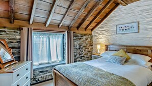 A bedroom with a stone wall, wooden ceiling beams, a bed with pillows, a bedside table with a lamp, and a window with curtains. - 1 Bed in Tavistock (oc-round) (Lamerton)