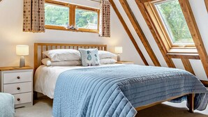 A bedroom with a sloped ceiling, a bed with a blue quilt, two bedside lamps, a nightstand, and curtains. - 3 Bed in Camelford (oc-pendw) (Davidstow)