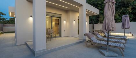 Un espacio lounge moderno al aire libre con sillones de sol y sombrillas, un acceso por puerta de cristal e iluminación ambiental.