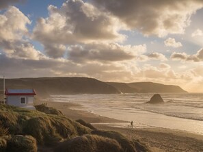 A coastal landscape with a small white building with a red roof, a grassy foreground, and a rocky outcrop in the sea. - 2 Bed in Beaworthy (oc-tilli) (Virginstowe)