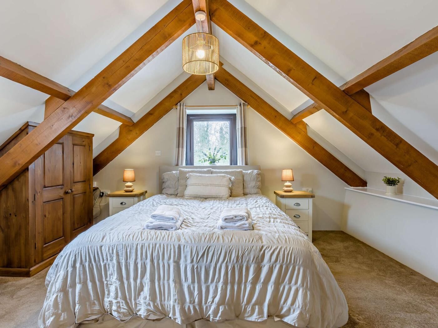 Un dormitorio con techo inclinado, vigas de madera, una cama con ropa de cama blanca, dos mesitas de noche con lámparas y una ventana con vistas a la vegetación.