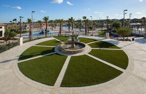 Ein kreisförmiger Platz mit einer zentralen Fontäne, umgeben von gepflegtem Rasen und einem Poolbereich im Hintergrund.