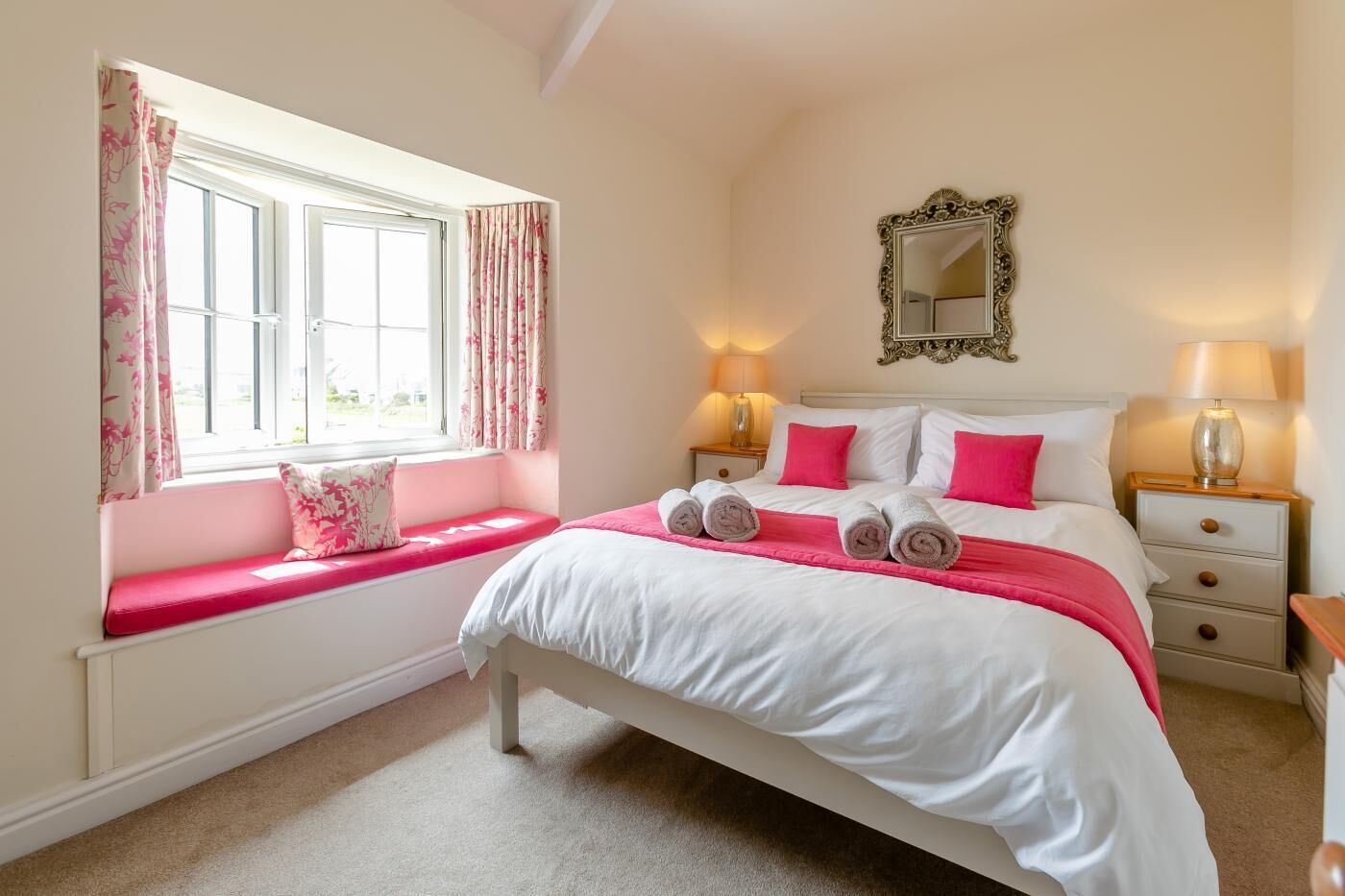 A bedroom with a bed, two bedside tables, a window with curtains, a mirror, and a bench with a cushion.