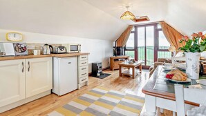 A kitchen with a refrigerator, microwave, and wooden countertop. There is a dining area with a table and chairs, and a view of the outdoors through a large window. - 1 Bed in Looe (oc-couvw) (Lansallos)