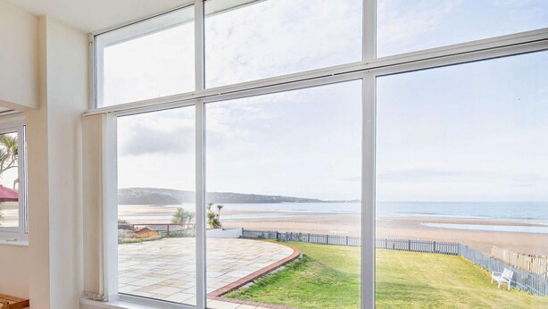 Ein Zimmer mit großen Fenstern und Blick auf einen Strand sowie einen eingezäunten Garten.
