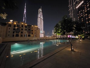 A nighttime pool area with a view of a tall building and another building with illuminated windows. - Gogoto7 Luxe 2BR Burj Khalifa View Prime Location (Dubai)