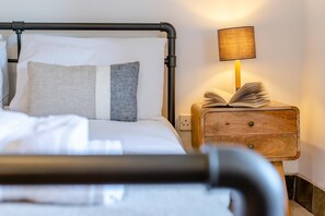 Ein Bett mit Metallgestell, weißer Bettwäsche und zwei Kissen. Ein Nachttisch aus Holz mit einer Lampe und einem offenen Buch steht daneben.