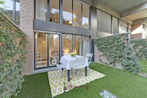 A modern building with large windows, a patio with a table and chairs, and a brick wall with climbing plants. - Garden Apartment in Center - Ambra (Lugano)
