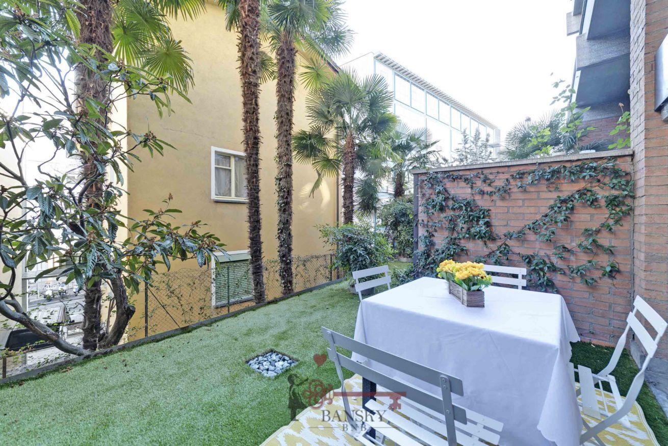 Un patio con una mesa y sillas, rodeado de plantas y un muro de ladrillo.