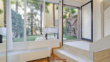 Una habitación con amplios ventanales, una mesa blanca con un florero y vistas a un jardín con palmeras.