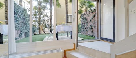 A room with large windows, a white table with a vase, and a view of a garden with palm trees.