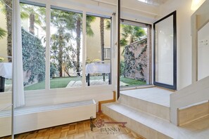 Quarto com grandes janelas, uma mesa branca com um vaso e vista para um jardim com palmeiras.