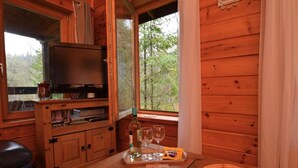 Interior de una cabaña de madera con televisión, una mesa con vasos y aperitivos, y una ventana con vista al bosque.