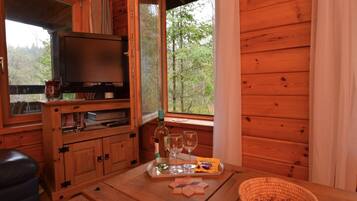 L’intérieur d’un chalet en bois, avec un téléviseur, une table avec des verres et des collations, et une fenêtre donnant sur la forêt.