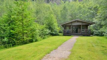 Un chalet rustique doté d’un porche, entouré d’une végétation luxuriante et accessible par un chemin de gravier.