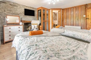 A bedroom with a stone wall, wooden paneling, a bed with floral bedding, a dresser, and a television. - 2 Bed in Liskeard (oc-04708) (Menheniot)