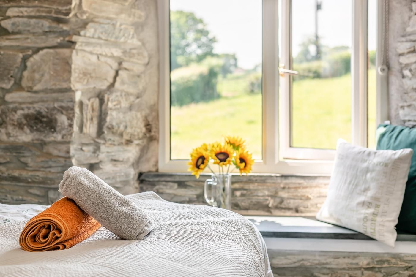 A room with a stone wall, a bed with a white blanket and an orange towel, a window with a view of greenery, and a vase with sunflowers.