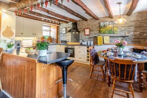 A rustic kitchen with wooden beams, a stone wall, and a dining area with a round table and chairs. - 2 Bed in Liskeard (oc-04708) (Menheniot)