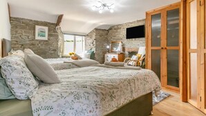 Une chambre avec un mur en pierre, un lit avec une literie à motifs floraux, une armoire en bois et un grand miroir.