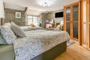 A bedroom with a stone wall, a bed with floral bedding, a wooden wardrobe, and a large mirror. - 2 Bed in Liskeard (oc-04708) (Menheniot)
