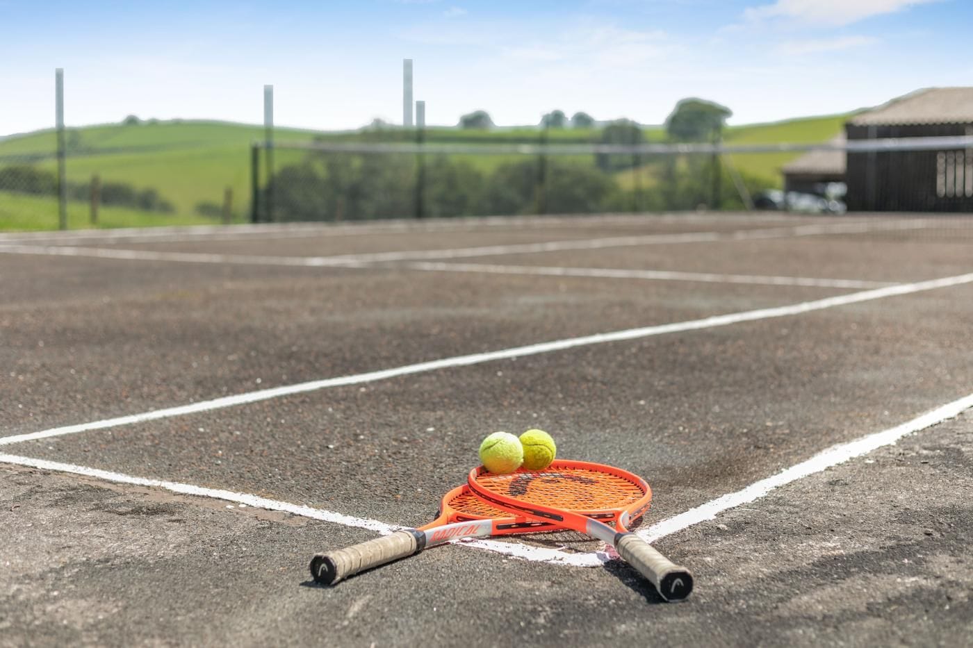 A tennis court with a racket and balls, surrounded by a fence and greenery.