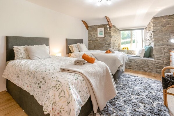 A bedroom with two beds, a stone wall, a window with a view, and a wooden floor. - 2 Bed in Liskeard (oc-04708) (Menheniot)