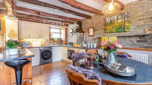 Une cuisine rustique avec un mur en pierre, des poutres en bois, un coin bar et une table à manger agrémentée d’un vase de fleurs.
