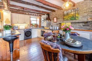 A rustic kitchen with a stone wall, wooden beams, a bar area, and a dining table with a vase of flowers. - 2 Bed in Liskeard (oc-04708) (Menheniot)
