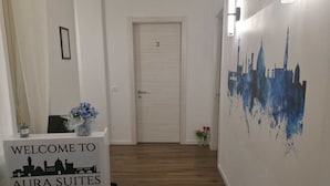 A hallway with a reception desk labeled 'Welcome to Aura Suites Florence,' a door marked with the number 3, and a wall mural of a city skyline.