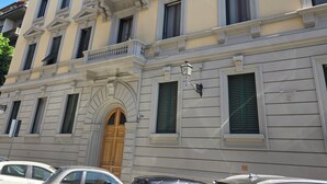 A multi-story building with a classic architectural style, featuring a central entrance with a wooden door, multiple windows with shutters, and a balcony with ornate railings.