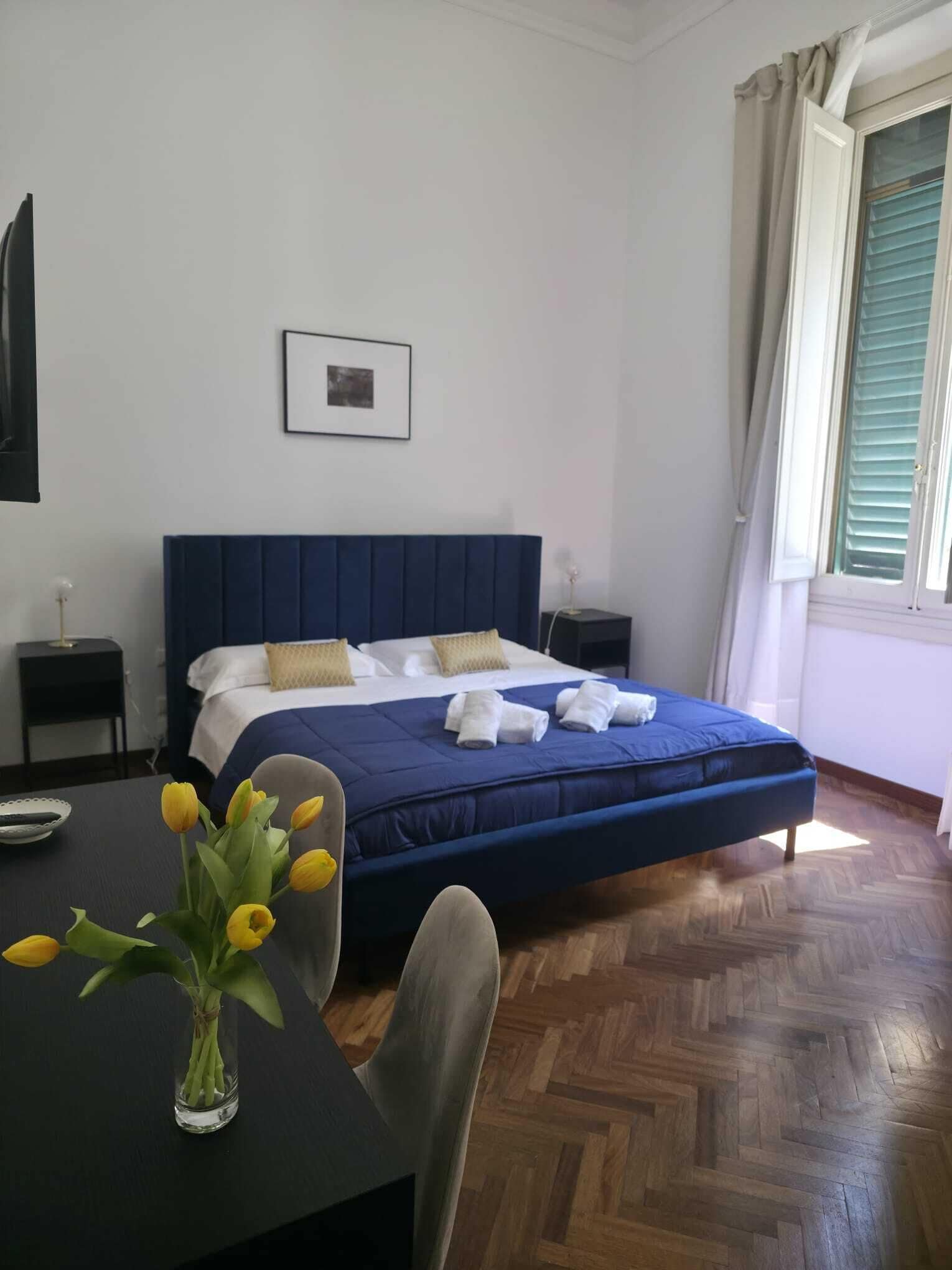 Quarto de hotel com cama, mesa de cabeceira, cadeira, escrivaninha com um vaso de flores e janela com cortinas.