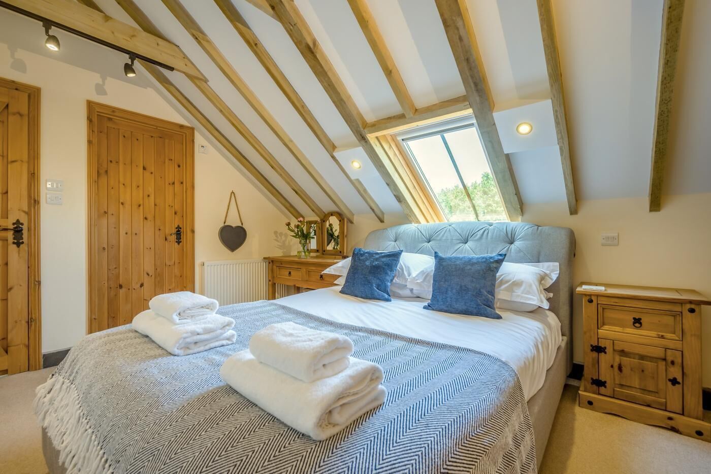 A bedroom with a large bed, wooden beams, a window, a dresser, and a hanging heart-shaped decoration.