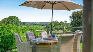 A wicker outdoor dining set with an umbrella, surrounded by greenery and a view of the countryside. - 3 Bed in Bude (oc-woott) (Poundstock)